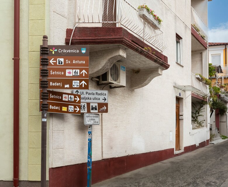 Ein Wegweiser an einer Straßenecke in einer ruhigen Stadt. Die Schilder zeigen verschiedene Richtungen zu Sehenswürdigkeiten und Straßen, während die umliegenden Gebäude eine charmante, mediterrane Architektur aufweisen.
