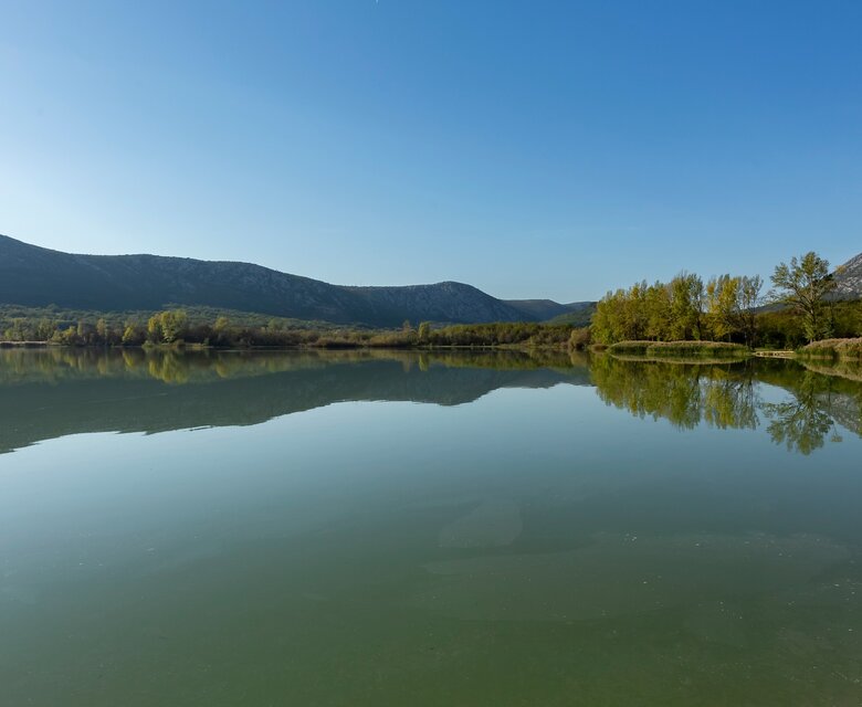 Ruhiger See mit spiegelndem Wasser, umgeben von sanften Hügeln und Bäumen. Der klare blaue Himmel reflektiert sich im Wasser, während die Landschaft eine friedliche Atmosphäre ausstrahlt.