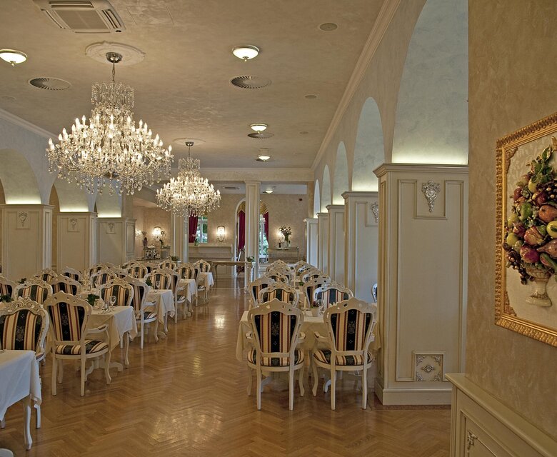 Elegantes Restaurant mit hellen Wänden und großen Kronleuchtern. Tische sind mit weißen Tischdecken gedeckt und Holzstühle stehen bereit. An den Wänden sind kunstvolle Dekorationen und ein Gemälde mit Früchten zu sehen. Die Atmosphäre ist stilvoll und einladend.