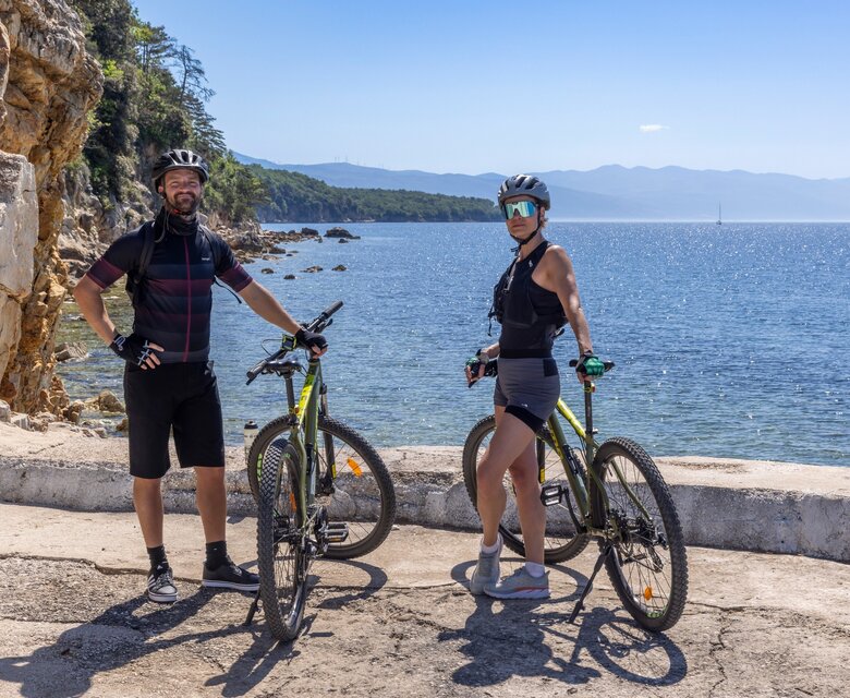 Zwei Radfahrer stehen mit ihren Mountainbikes an einer Küstenstraße. Im Hintergrund erstreckt sich das glitzernde Wasser des Meeres und eine malerische Landschaft mit Bergen. Die Sonne scheint, und der Himmel ist klar und blau.