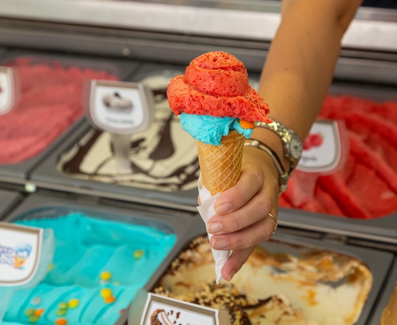Eine Hand hält ein Waffeleis mit zwei Kugeln: eine rote und eine türkisfarbene. Im Hintergrund sind verschiedene Eissorten in Behältern sichtbar, die eine bunte Auswahl an Geschmäckern zeigen.