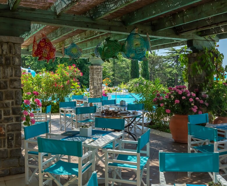 Ein einladender Außenbereich eines Restaurants mit weißen Tischen und türkisfarbenen Stühlen, umgeben von üppigen Pflanzen und bunten Blumen. Im Hintergrund ist ein Pool sichtbar, der eine entspannte Atmosphäre schafft. Farbige Laternen hängen von der Überdachung.