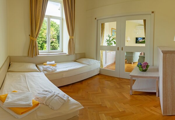 Helles Hotelzimmer mit zwei Betten, einem Tisch und einer Pflanze. Große Fenster lassen Tageslicht herein und bieten Blick ins Grüne. Der Boden ist aus Holz, und die Wände sind in sanften Farben ge... Hotel mit Appartements für Familien, Kroatien - am Meer