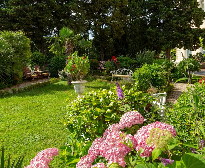 Ein blühender Garten mit bunten Blumen, grünen Sträuchern und Bäumen. Im Hintergrund sind gemütliche Sitzgelegenheiten und ein Pavillon sichtbar. Die Atmosphäre ist einladend und friedlich, ideal für Entspannung im Freien.