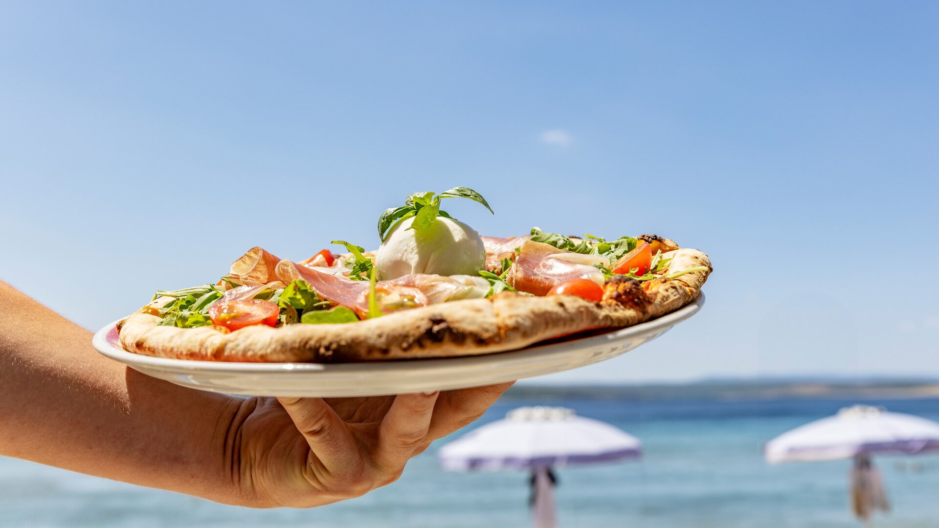 Eine Hand hält einen Teller mit einer frisch zubereiteten Pizza, belegt mit Schinken, Tomaten und Rucola. Im Hintergrund ist ein schöner Strand mit Sonnenschirmen und klarem Wasser zu sehen, unter blauem Himmel.