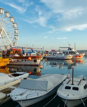 Hafen mit verschiedenen Booten, darunter ein gelbes Boot und ein weißes Motorboot, die sanft im Wasser schaukeln. Im Hintergrund ist ein Riesenrad zu sehen, unter einem klaren blauen Himmel mit wenigen Wolken.