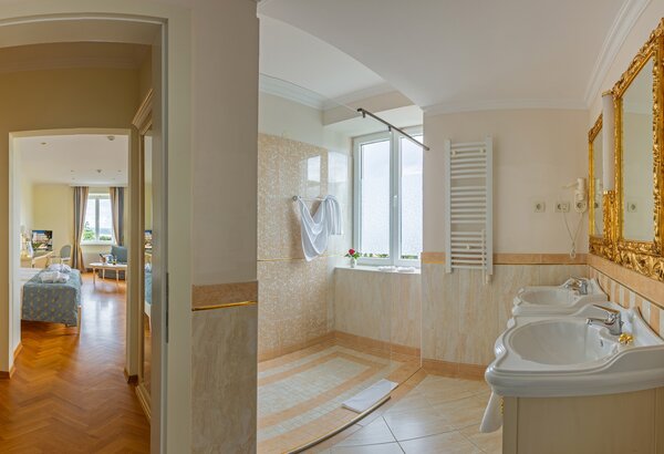 Helles Badezimmer mit zwei Waschbecken, großen Spiegeln und einem Fenster, das Tageslicht hereinlässt. Im Hintergrund ist ein gemütliches Schlafzimmer mit einem Bett und einem Fernseher sichtbar. D... 4-Sterne-Hotel am Meer Kroatien ☛ Superior Doppelzimmer