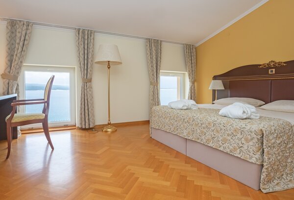 Helles Hotelzimmer mit einem großen Doppelbett, zwei Fenstern mit Blick auf das Wasser und einem Schreibtisch mit Stuhl. Die Wände sind in warmen Farben gehalten, und der Boden ist aus Holz. Meerblick Hotel Kroatien - Ihr Komfort Doppelzimmer