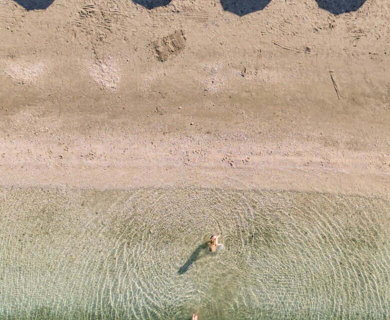 Ein Blick von oben auf einen Strand mit Sonnenschirmen und Liegen. Im klaren Wasser sind drei Personen, die schwimmen. Die sanften Wellen erzeugen kreisförmige Muster, während der Sandstrand im Hintergrund sichtbar ist.