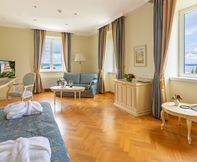 Helles Hotelzimmer mit Holzfußboden, großen Fenstern und Blick auf die Landschaft. Es gibt ein bequemes Sofa, einen Tisch mit Blumen und einen Fernseher. Die Wände sind in sanften Farben gehalten, und die Vorhänge sind elegant drapiert.