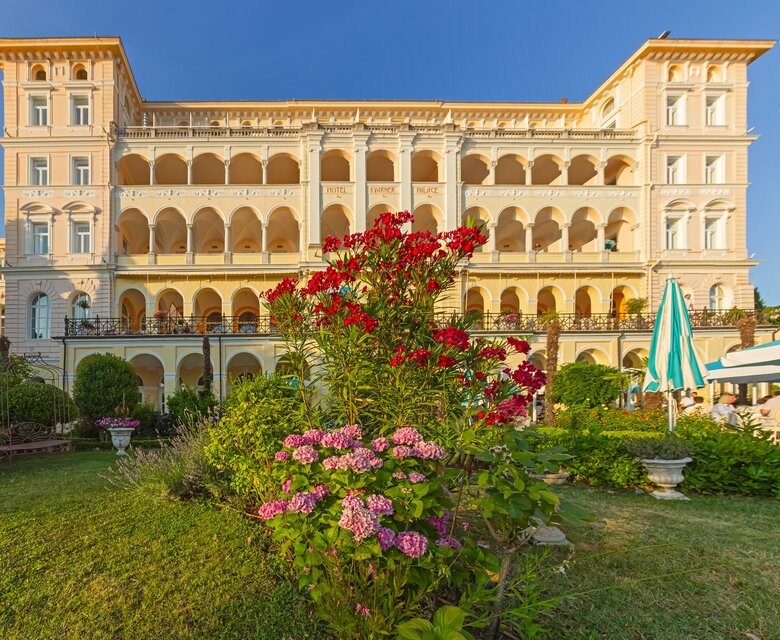Ein elegantes Hotelgebäude mit mehreren Etagen und Säulen, umgeben von einem gepflegten Garten mit bunten Blumen. Im Vordergrund blühen rosa und rote Blumen, während im Hintergrund Sonnenschirme und Sitzbereiche für Gäste sichtbar sind. Der Himmel ist klar und blau.