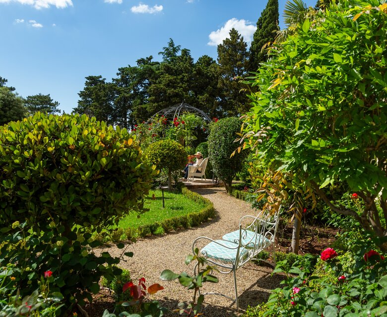 Ein malerischer Garten mit üppigem Grün, bunten Blumen und einem geschwungenen Kiesweg. Im Hintergrund sitzt eine Person auf einer Bank und genießt die ruhige Atmosphäre unter blauem Himmel und vereinzelten Wolken.