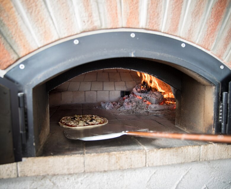 Ein Holzofen mit flammendem Feuer, in dem eine Pizza auf einem Schaufelbrett liegt. Die Backfläche ist aus Stein, und die Atmosphäre ist warm und einladend.