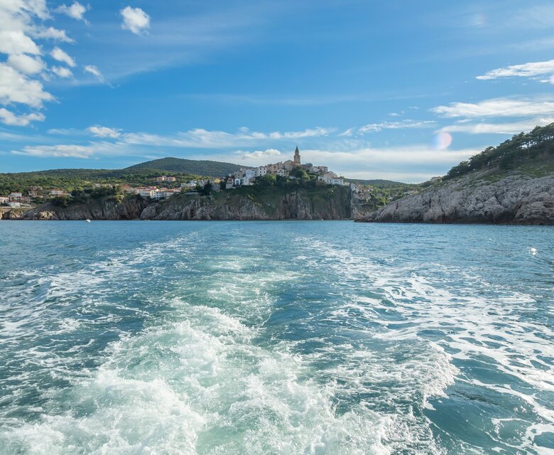 Blick auf eine malerische Küstenstadt, die auf einem Hügel thront, während sich das Boot durch das klare Wasser bewegt. Im Hintergrund sind grüne Hügel und ein blauer Himmel mit einigen Wolken zu sehen.