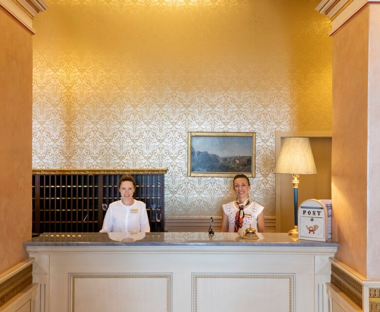Zwei Empfangsdamen stehen an der Rezeption eines eleganten Hotels mit goldenen Wänden und stilvollen Lampen. Im Hintergrund sind Postfächer und ein Gemälde sichtbar, was eine einladende Atmosphäre schafft.
