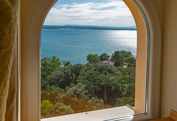 Blick aus einem großen, bogenförmigen Fenster auf einen ruhigen See, umgeben von üppigem Grün und Bäumen. Die Sonne reflektiert auf der Wasseroberfläche, während sanfte Wolken den blauen Himmel sch... Appartements für Familienurlaub Crikvenica, Kroatien