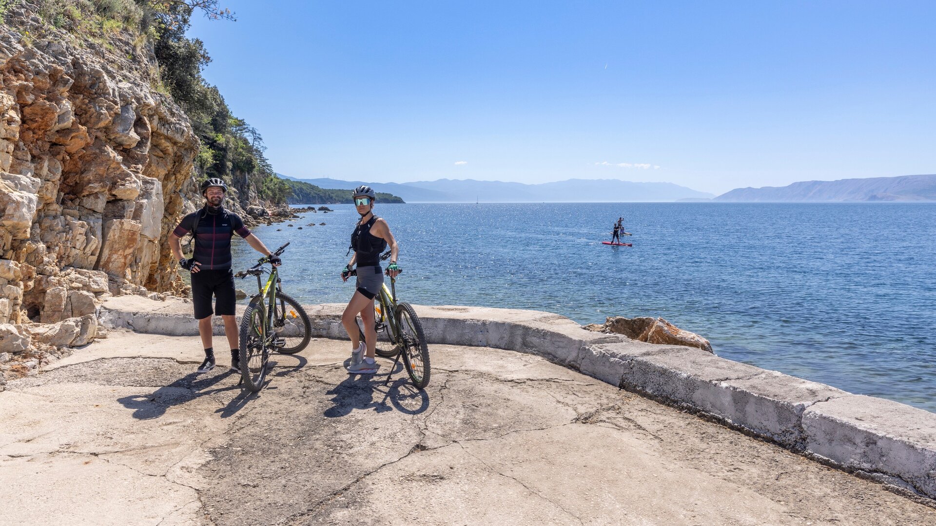 Zwei Radfahrer stehen auf einem gepflasterten Weg am Wasser, umgeben von Felsen und Bäumen. Im Hintergrund ist ein ruhiger See mit einem Paddler zu sehen. Der Himmel ist klar und sonnig, was eine entspannte Atmosphäre schafft.