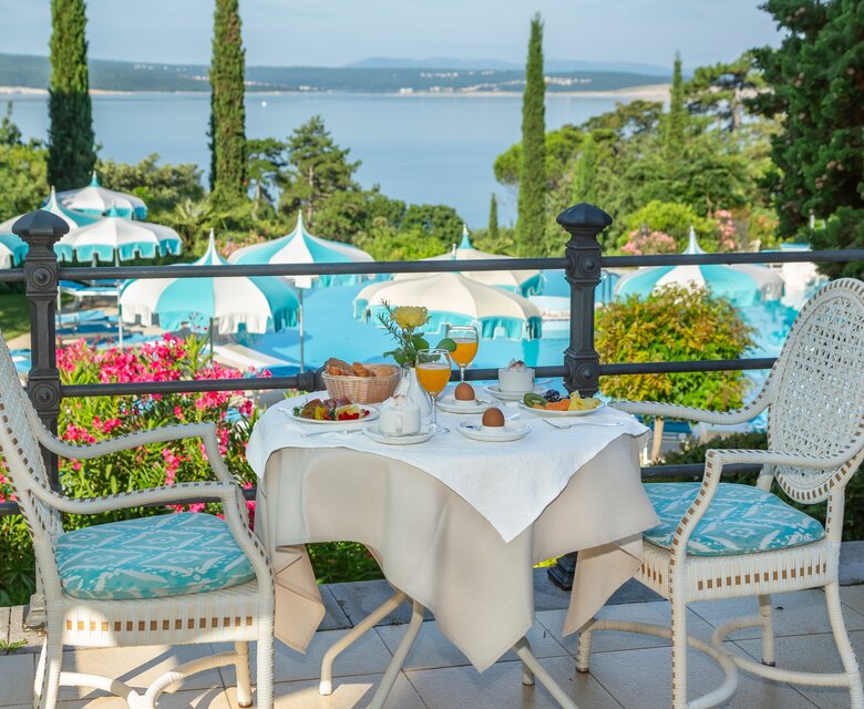 Ein gedeckter Tisch mit Frühstück auf einer Terrasse, umgeben von blühenden Pflanzen und Blick auf einen Pool und den See im Hintergrund. Sonnige Atmosphäre mit grünen Bäumen und blauen Sonnenschirmen.