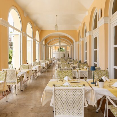 Heller Speisesaal mit großen Fenstern und eleganten Tischen, die mit gelben Tischdecken und weißen Servietten gedeckt sind. Die Wände sind in sanften Gelbtönen gehalten, und die Atmosphäre wirkt einladend und freundlich.