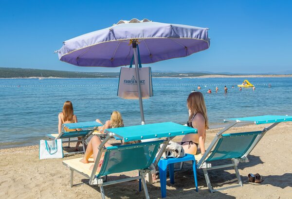 Drei Frauen entspannen unter einem Sonnenschirm am Strand, während sie auf Liegen sitzen. Im Hintergrund ist das ruhige Wasser und einige Badegäste zu sehen, die im Meer schwimmen. Der Himmel ist klar und sonnig.