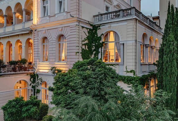 Eine elegante Hotelansicht mit hell erleuchteten Fenstern, umgeben von üppigem Grün und Bäumen. Die Architektur ist klassisch mit großen Fenstern und Balkonen, die eine einladende Atmosphäre schaffen.