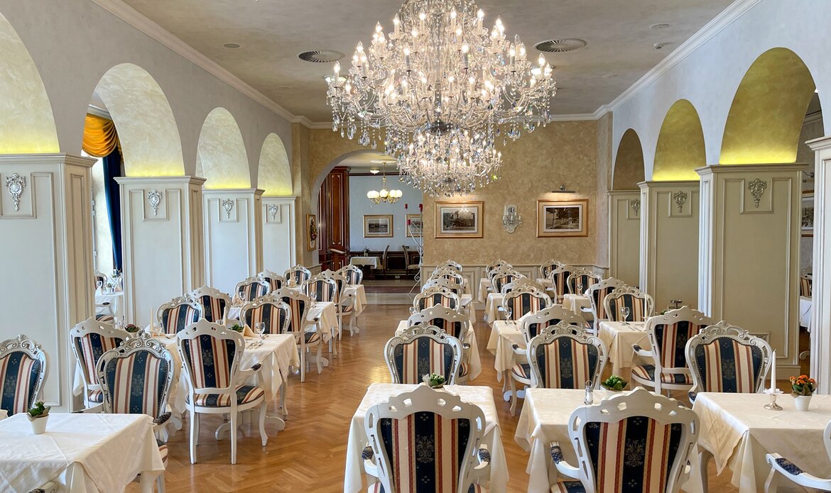 Elegantes Restaurant mit weißen Tischen und gestreiften Stühlen. Ein großer Kristalllüster hängt von der Decke, während große Fenster für Tageslicht sorgen. Die Wände sind in sanften Farben gehalten, und der Raum strahlt eine gehobene Atmosphäre aus.