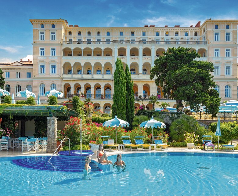 Ein luxuriöses Hotel mit einer eleganten Fassade steht im Hintergrund, während im Vordergrund ein einladender Pool mit Sonnenliegen und Sonnenschirmen zu sehen ist. Einige Gäste genießen das Wasser, umgeben von üppigen Gärten und blühenden Pflanzen.