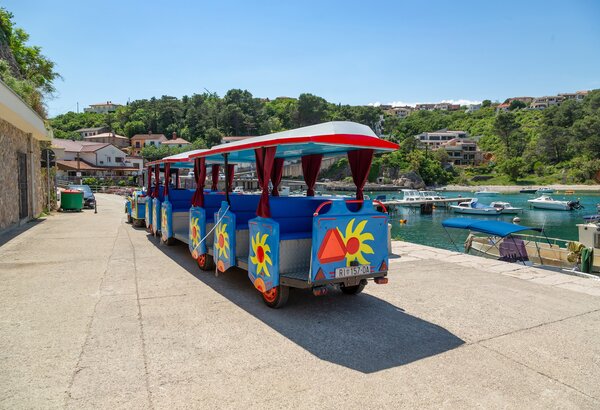 Bunte Touristenbahn auf einem gepflasterten Weg am Hafen, umgeben von grünen Hügeln und kleinen Booten im Wasser. Sonniger Himmel und eine entspannte Urlaubsatmosphäre.