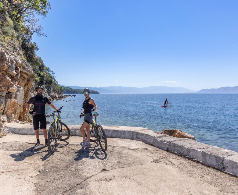 Zwei Radfahrer stehen auf einem Uferweg am Wasser, umgeben von Felsen und grüner Vegetation. Im Hintergrund ist ein ruhiger See und ein weiterer Wassersportler auf einem Stand-Up-Paddle-Board zu sehen. Der Himmel ist klar und sonnig.