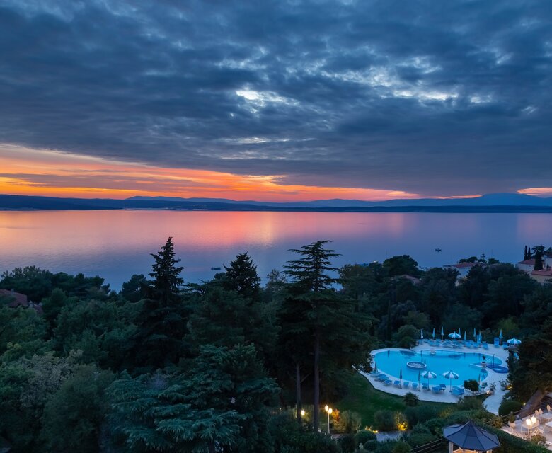 Blick auf einen ruhigen See bei Sonnenuntergang, umgeben von Bäumen und einem einladenden Poolbereich. Die Wolken reflektieren die warmen Farben des Himmels, während die Landschaft eine friedliche Atmosphäre ausstrahlt.