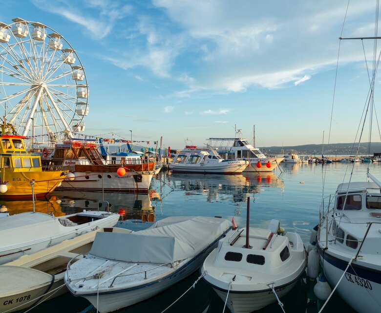 Blick auf einen Hafen mit verschiedenen Booten und einem Riesenrad im Hintergrund. Die Wasseroberfläche spiegelt die Farben des Himmels wider, während die Atmosphäre ruhig und einladend wirkt.