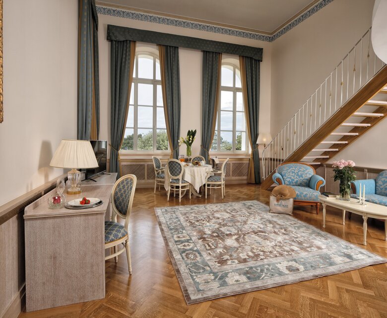 Geräumiges Hotelzimmer mit eleganter Einrichtung, großen Fenstern und einem gemütlichen Sitzbereich. Ein Tisch mit Stühlen steht neben einer Treppe, während ein Teppich den Boden schmückt. Pflanzen und Dekoration verleihen dem Raum eine einladende Atmosphäre.