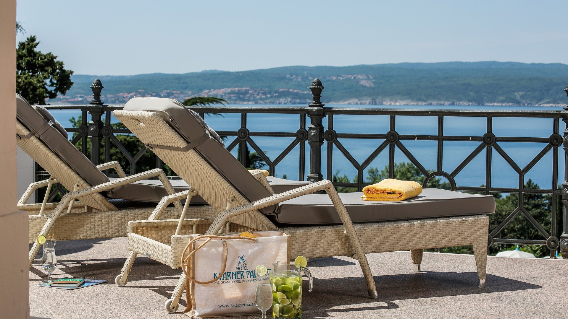 Zwei Liegen stehen auf einer Terrasse mit Blick auf das Wasser. Eine gelbe Handtuch liegt auf einer Liege, während eine Tasche mit Getränken und frischen Früchten daneben steht. Die Umgebung strahlt eine entspannte Urlaubsatmosphäre aus.