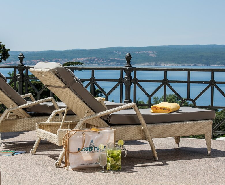 Zwei Liegen stehen auf einer Terrasse mit Blick auf das Wasser. Eine gelbe Handtuch liegt auf einer Liege, während eine Tasche mit Getränken und frischen Früchten daneben steht. Die Umgebung strahlt eine entspannte Urlaubsatmosphäre aus.