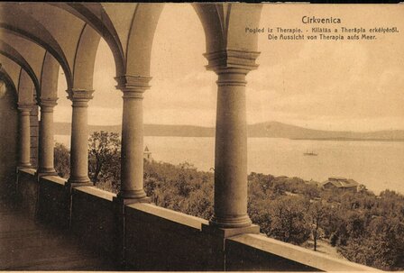 Blick auf das Meer von einer überdachten Terrasse mit Säulen. Im Hintergrund sind Bäume und Gebäude zu sehen, während ein Schiff auf dem Wasser fährt. Die Atmosphäre ist ruhig und einladend.