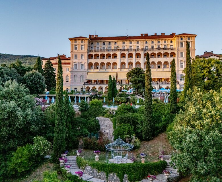 Ein elegantes Hotelgebäude umgeben von üppigem Grün und Bäumen. Im Vordergrund befindet sich ein gepflegter Garten mit einem Pavillon und bunten Blumenbeeten. Die Atmosphäre ist ruhig und einladend, ideal für Erholung und Entspannung.
