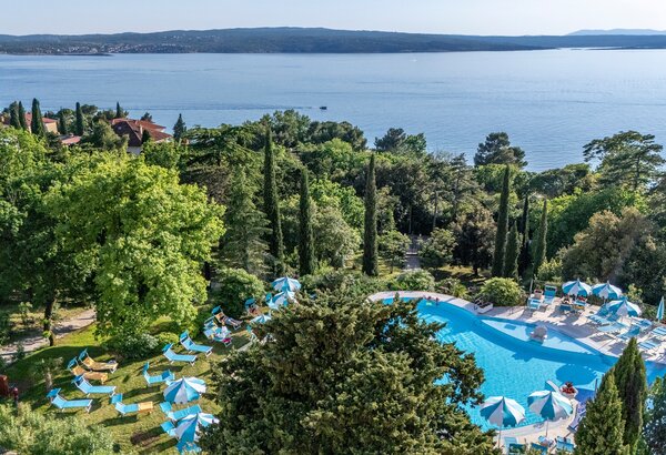 Ein Blick auf einen Poolbereich umgeben von üppigem Grün und Zypressen, mit Liegen und Sonnenschirmen. Im Hintergrund erstreckt sich das ruhige Wasser eines Sees und die sanfte Hügellandschaft.