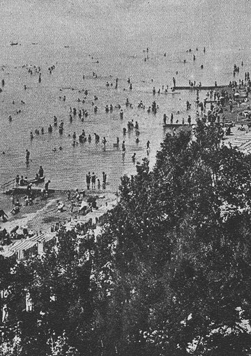 Ein historisches Schwarz-Weiß-Bild eines Strandes mit vielen Menschen, die im Wasser schwimmen oder am Ufer entspannen. Im Vordergrund sind Liegen und Sonnenschirme zu sehen, während im Hintergrund das Meer und einige Boote sichtbar sind.
