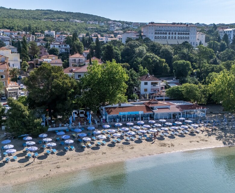 Ein Blick auf einen Strand mit vielen Sonnenschirmen und Liegen, umgeben von Bäumen. Im Hintergrund sind mehrere Gebäude und Hügel zu sehen, die eine entspannte Urlaubsatmosphäre schaffen. Das Wasser ist ruhig und klar.