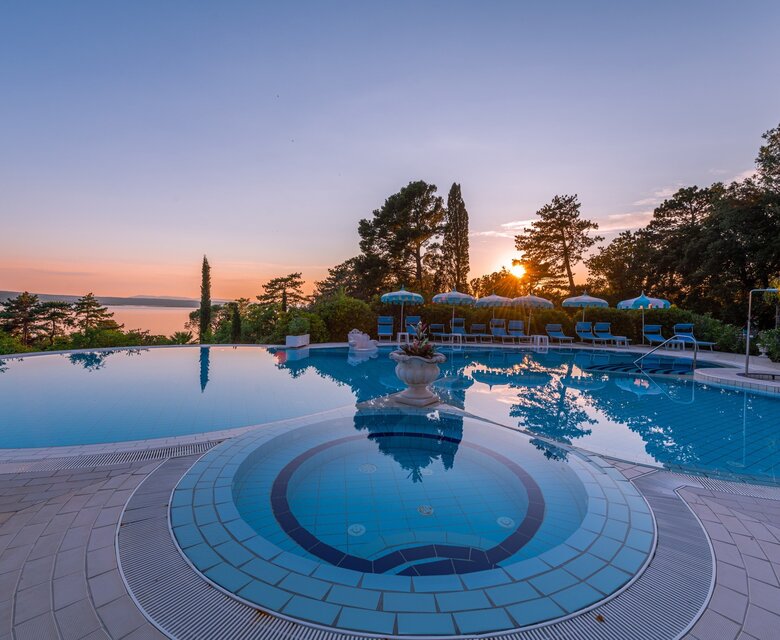 Ein schöner Pool mit einem runden Bereich im Vordergrund, umgeben von Liegen und Sonnenschirmen. Im Hintergrund ist ein Sonnenuntergang über dem Wasser zu sehen, umgeben von Bäumen und einer ruhigen Atmosphäre.