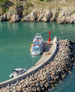Ein Boot liegt an einem Steg in einer ruhigen, türkisfarbenen Bucht. Im Hintergrund sind steile Felsen zu sehen. Ein rotes Leuchtfeuer markiert den Hafen, während ein weiteres kleines Boot in der Nähe ankert. Die Atmosphäre ist friedlich und einladend.