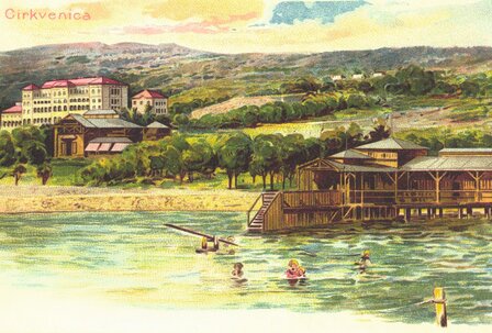 Blick auf einen malerischen See mit einem Holzsteg und Schwimmern. Im Hintergrund sind grüne Hügel und historische Gebäude zu sehen, die eine entspannte Urlaubsatmosphäre vermitteln.