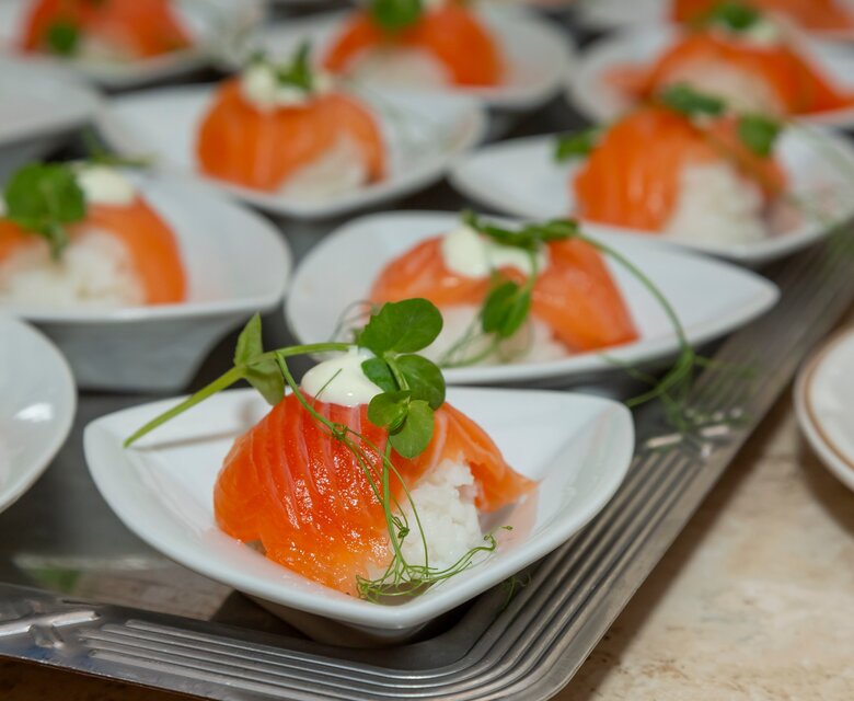 Eine Reihe von kleinen Schalen mit Sushi, gefüllt mit Reis und belegt mit frischem Lachs. Jede Portion ist mit einem Klecks Creme und frischen Kräutern garniert, auf einem silbernen Tablett angerichtet.