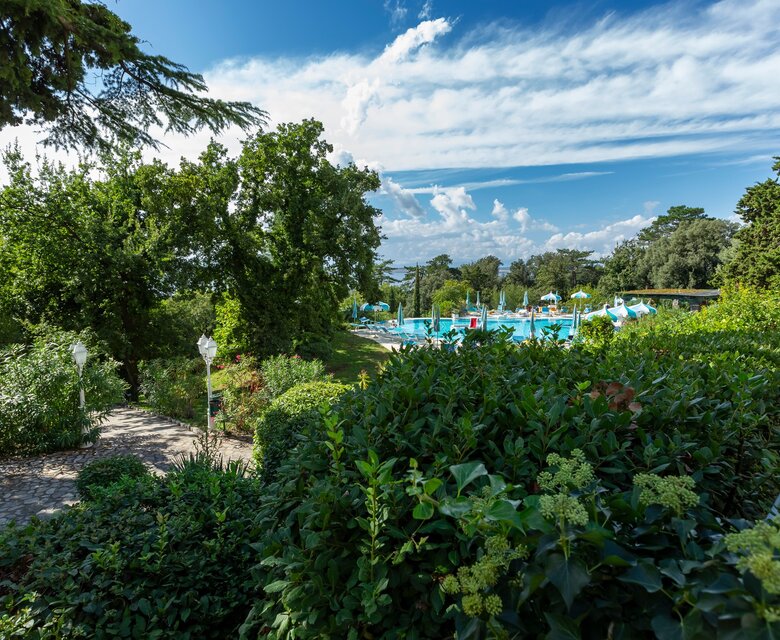 Ein Blick auf einen gepflegten Garten mit üppigem Grün und Bäumen, der zu einem einladenden Poolbereich führt. Der Himmel ist blau mit einigen Wolken, und der Pool ist mit bunten Liegen und Sonnenschirmen ausgestattet.