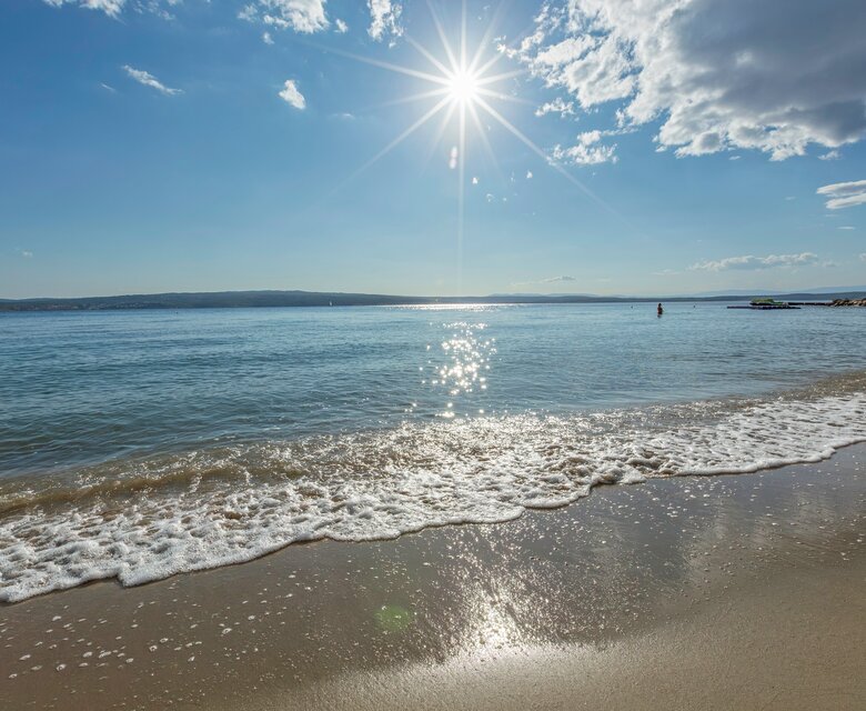 Ein sonniger Strand mit sanften Wellen, die an den Sand schlagen. Im Hintergrund erstreckt sich das ruhige Wasser bis zum Horizont, während die Sonne hell am Himmel strahlt und glitzernde Reflexionen auf der Wasseroberfläche erzeugt.
