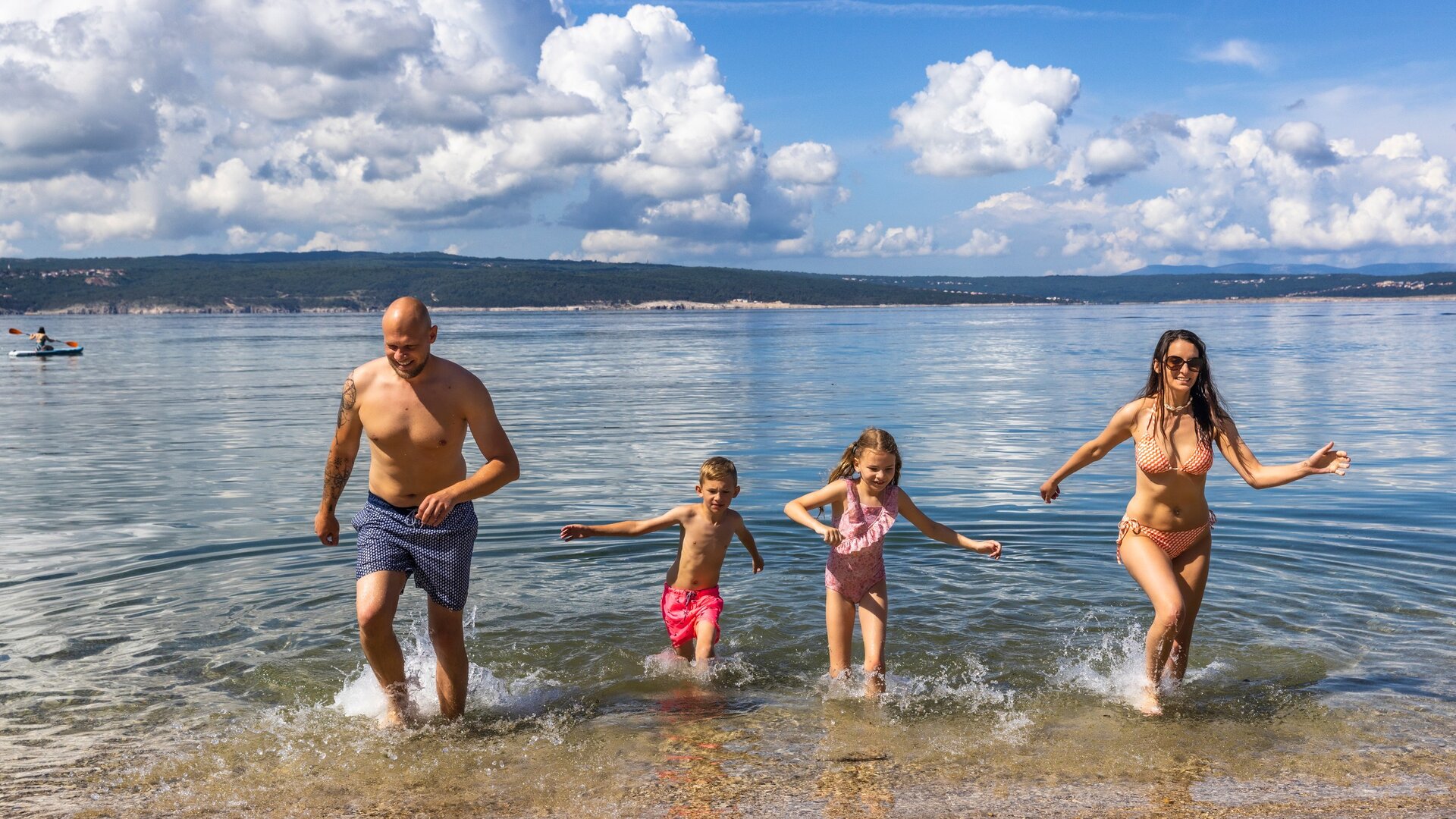 Eine Familie mit zwei Kindern läuft fröhlich ins Wasser eines klaren Sees. Die Erwachsenen tragen Badebekleidung, während die Kinder in bunten Schwimmanzügen sind. Im Hintergrund sind sanfte Wellen und eine malerische Landschaft mit Wolken zu sehen.