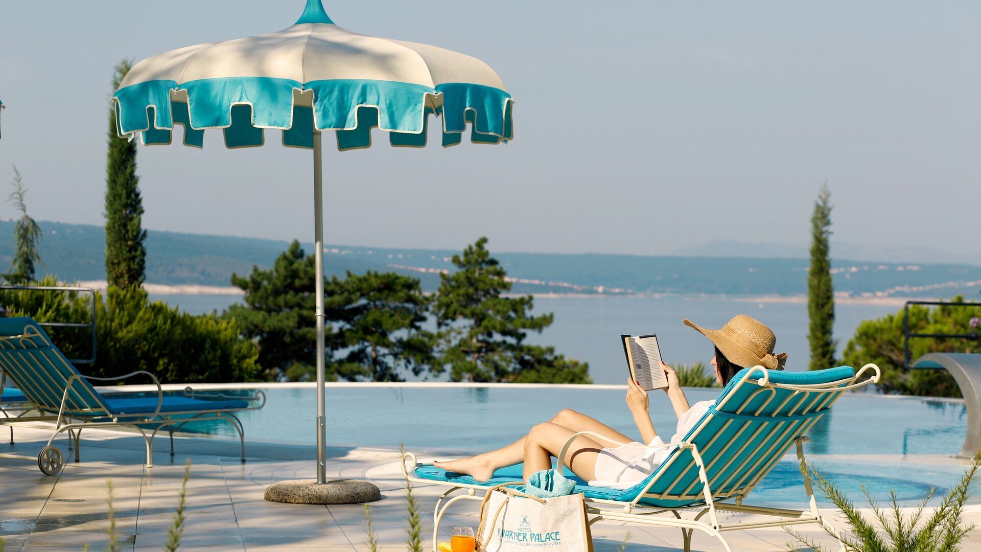 Eine Person entspannt auf einer Liege am Pool, liest ein Buch und trägt einen großen Sonnenhut. Im Hintergrund ist eine malerische Aussicht auf das Wasser und grüne Bäume zu sehen. Ein Sonnenschirm bietet Schatten.