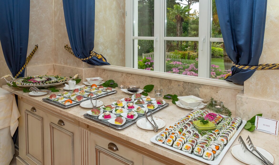 Ein eleganter Buffet-Tisch mit verschiedenen Sushi- und Fingerfood-Platten, dekoriert mit frischen Blumen und Blättern. Im Hintergrund sind große Fenster mit blauen Vorhängen, die einen Blick auf einen blühenden Garten bieten.