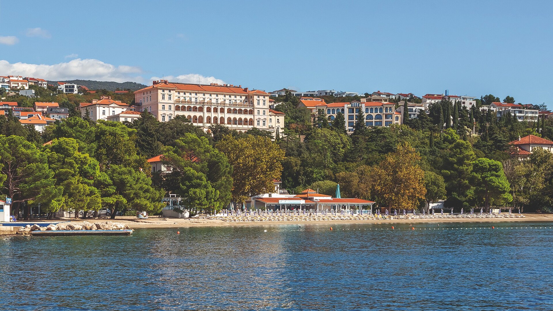 Blick auf eine Küstenlandschaft mit einem Hotelgebäude im Hintergrund, umgeben von Bäumen und bunten Häusern. Der Strand im Vordergrund ist mit Liegen ausgestattet, und das Wasser ist ruhig und klar. Der Himmel ist blau mit wenigen Wolken.