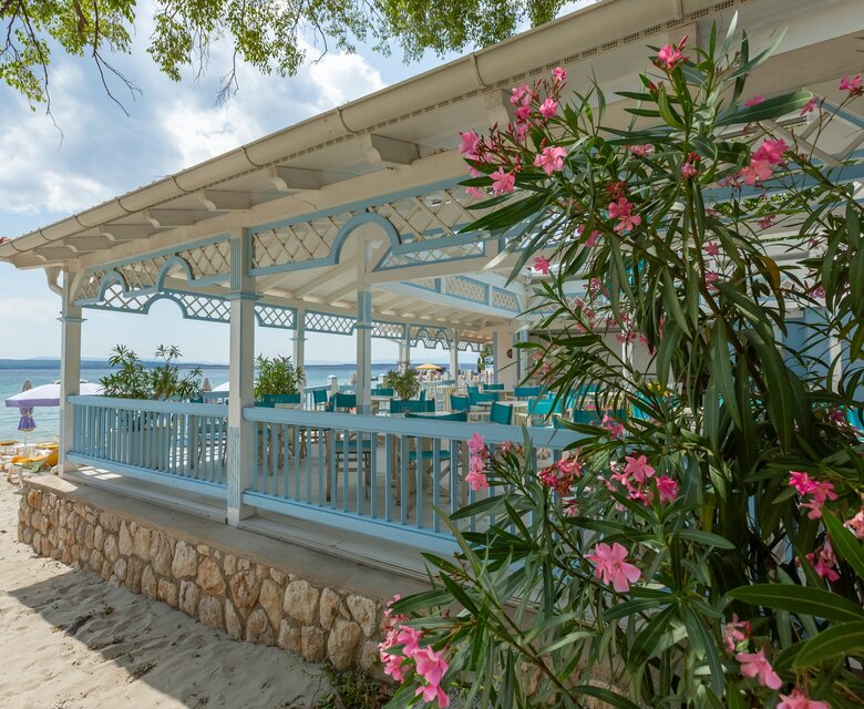 Ein einladender Strandpavillon mit blauen Möbeln und bunten Blumen im Vordergrund. Der Pavillon bietet einen Blick auf das ruhige Wasser und die umliegenden Sonnenliegen. Die Atmosphäre ist entspannt und tropisch, ideal für einen erholsamen Tag am Strand.